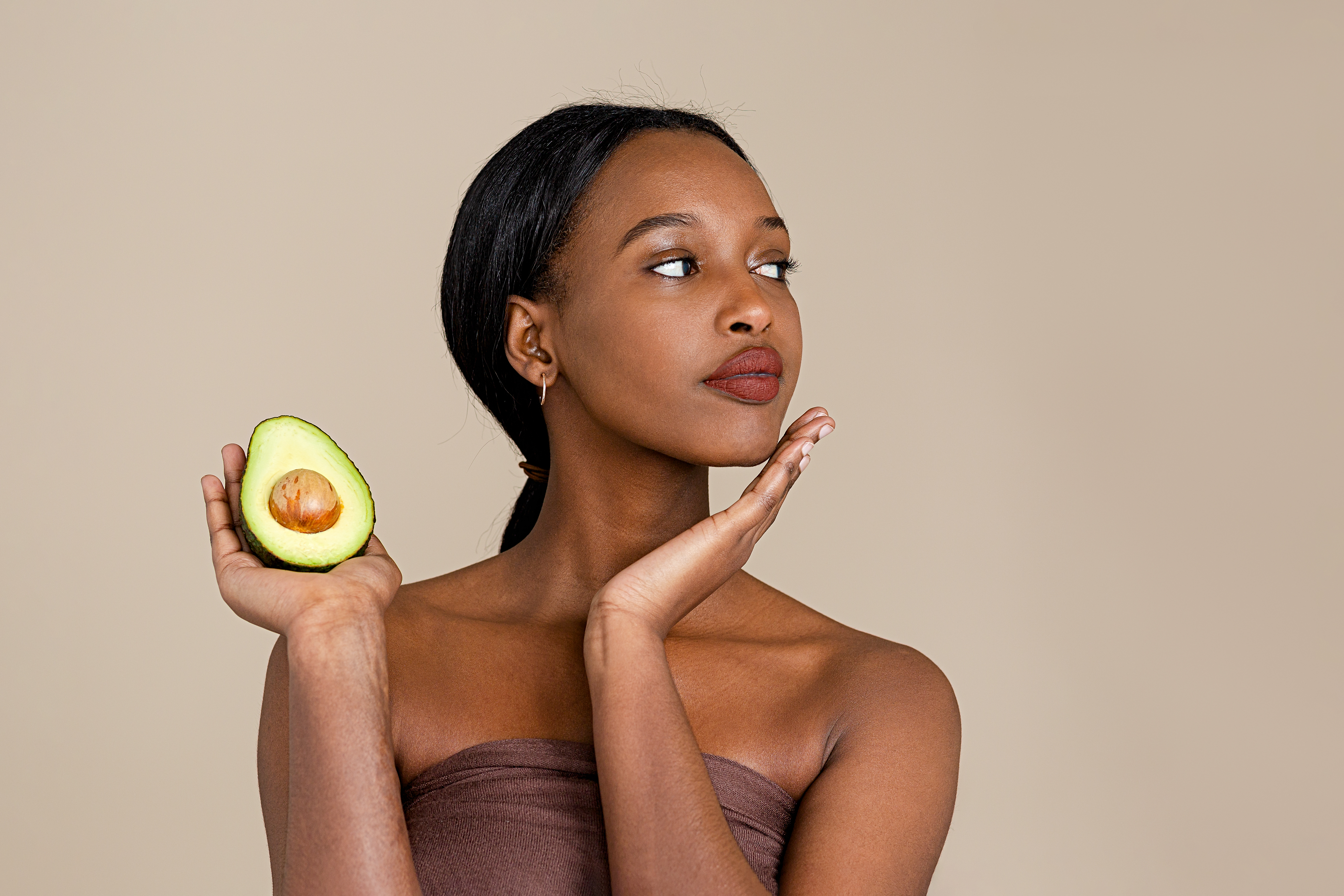 Natural beauty Mulher olhando para o lado com uma das mãos no queixo e a outra segurando metade de um avocado e usando toalha no corpo e com rabo de cavalo baixo