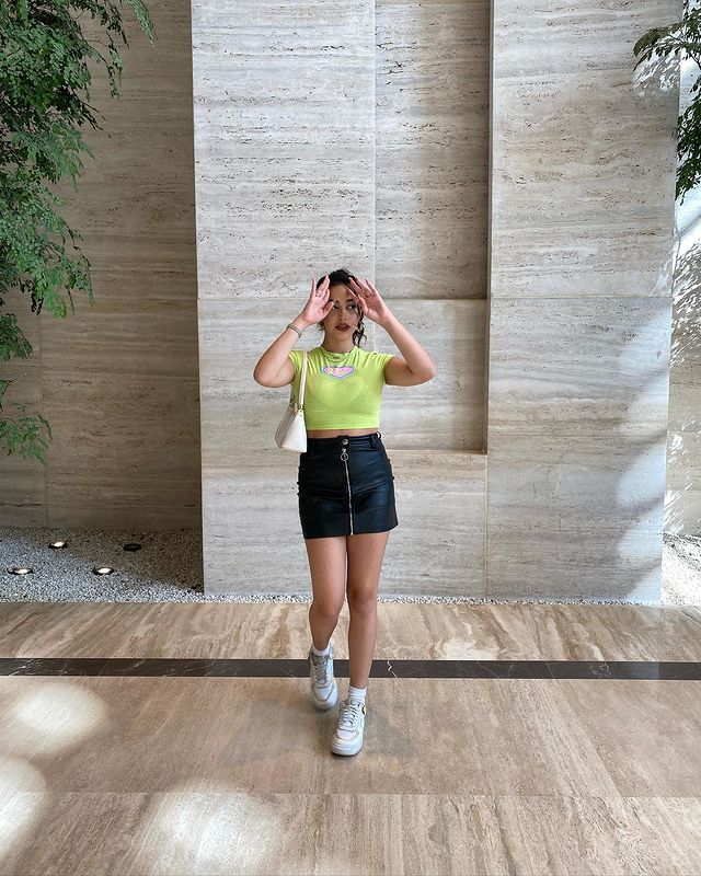 baby-tee-com-saia Foto de uma mulher. Ela usa uma baby tee verde, saia de couro preta, tênis branco, bolsa baguete branca e cabelo preso. Ela está com as duas mãos no cabelo, olha para o lado e não sorri.