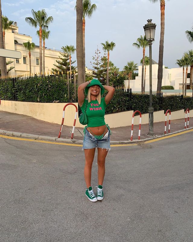 baby-tee-com-shorts Foto de uma mulher na rua. Ela usa um chapéu de pescador verde, baby tee verde, short jeans, bolsa verde e tênis verde. Ela está com as duas mãos na cabeça, de olho fechado e sorri para a foto.