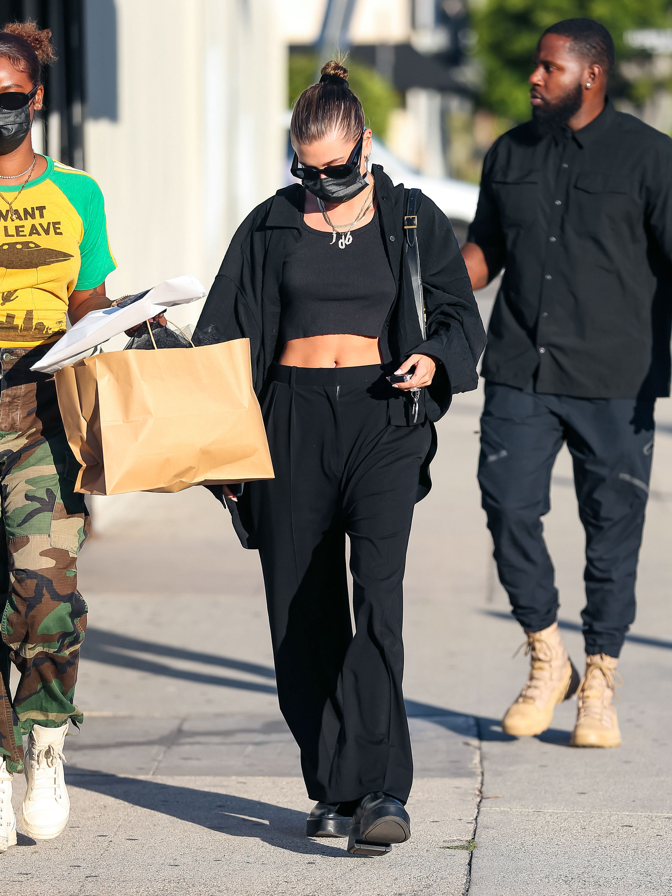 Hailey Bieber usando look todo preto com top cropped, calça de alfaiataria, camisa, tênis, bolsa pendurada no ombro, máscara de proteção facial e óculos escuros