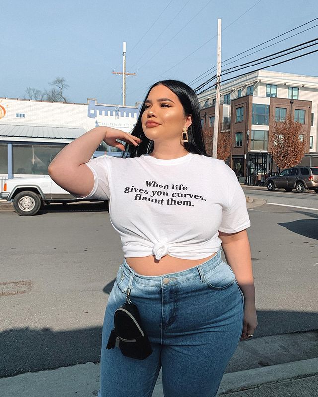 Fpto de uma mulher na rua. Ela usa uma camiseta branca com um nó frontal, calça jeans e bolsinha pendurada na calça. Ela está de cabelo solto, maquiagem glow e brinco dourado. Ela olha para o lado e sorri levemente.