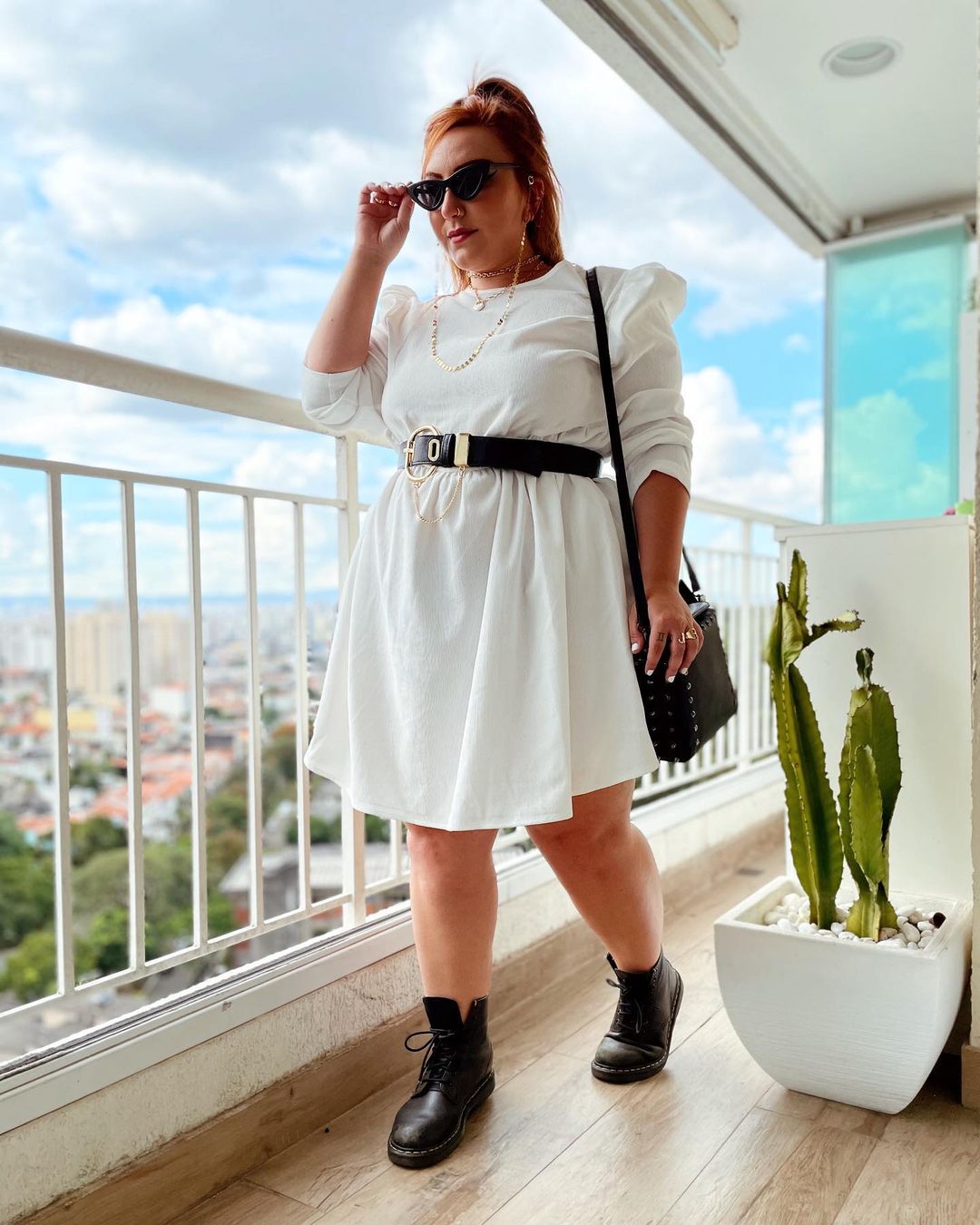look-vestido-branco-ju_romano Garota usando vestido branco com cinto preto, botinha preta, bolsa e óculos de sol pretos. Seu cabelo está preso em um rabo e ela está na varanda de um apartamento