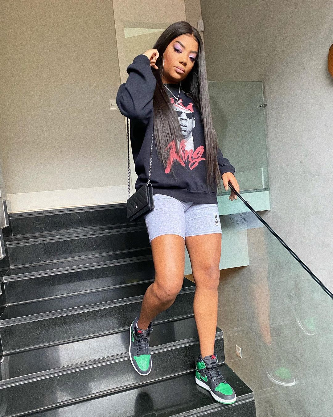 ludmilla-look-brinco-argola Ludmilla descendo escadas usando look com blusa de moletom preta, biker short cinza, tênis Nike verde e preto e bolsa preta. Ela está com uma das mãos no corrimão e a outra no cabelo.