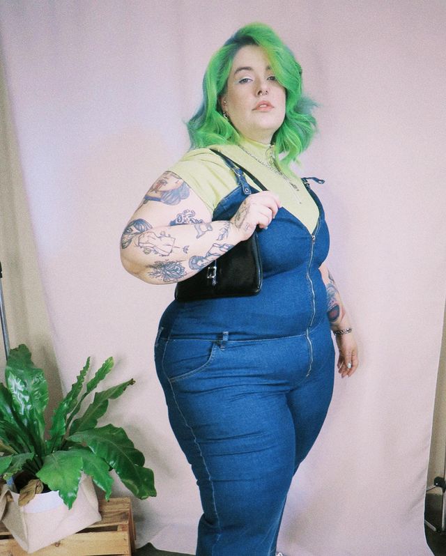 Foto de uma mulher. Ela usa um macacão jeans, camiseta verde e bolsa baguete preta. Ela olha para a câmera e não sorri.