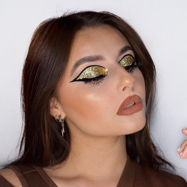 Foto do rosto de uma mulher. Ela usa uma blusa marrom, cabelo solto, brinco de argola pequeno e maquiagem com pele glow, batom marrom e olho com glitter dourado na pálpebra e delineado preto. Ela olha para o lado e não sorri para a foto.