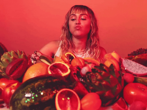 miley-cyrus-comendo-fruta Miley Cyrus em frente a uma pilha de frutas comendo fruta