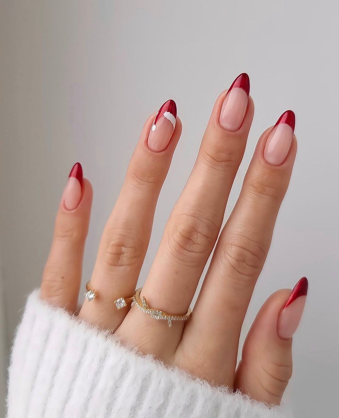 nail-art-natal-naillington Mão com unhas com francesinha vermelha de Natal, anéis dourados em dois dedos e parte de uma blusa branca de manga comprida