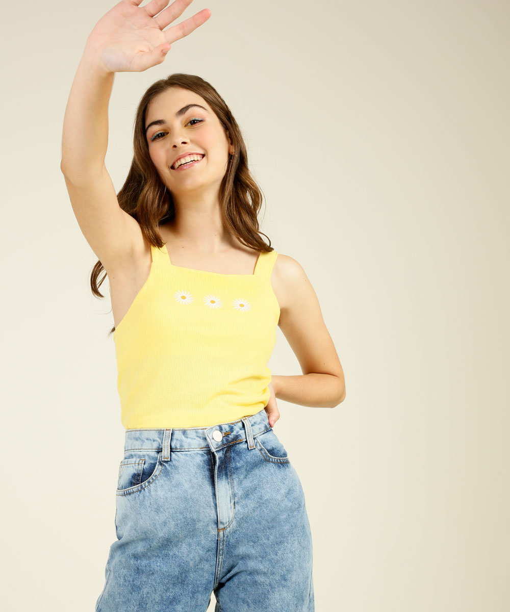 top-amarelo-colecao-capricho-marisa Garota usando top cropped amarelo com flores e calça jeans, sorrindo, com uma das mãos na cintura e a outra esticada para cima