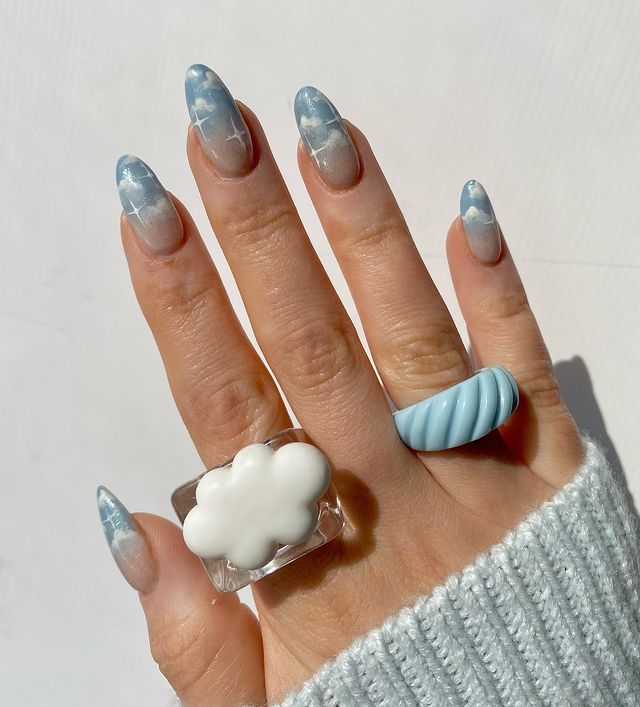 Foto de uma mão com anéis divertidos no dedo indicador e anelar com as unhas decoradas com unhas azuis e detalhe de nuvens brancas.