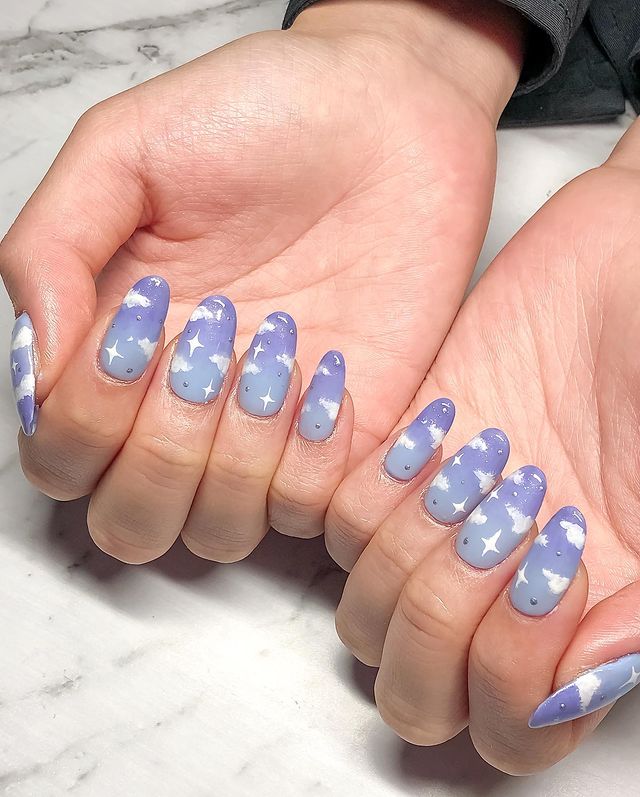 unhas-lilas-com-decoracao-de-nuvens Foto de mãos com as unhas decoradas com esmalte lilás e desenho de nuvens brancas.