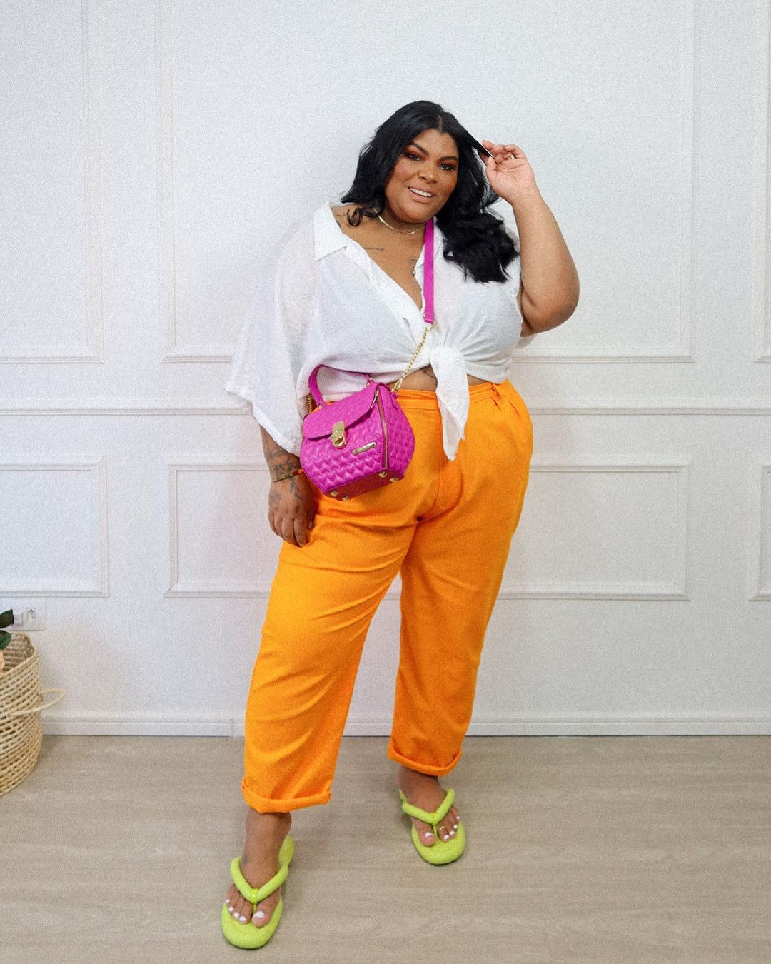 Mulher sorrindo com uma das mãos no cabelo usando camisa branca, calça laranja, bolsa rosa e chinelo amarelo neon