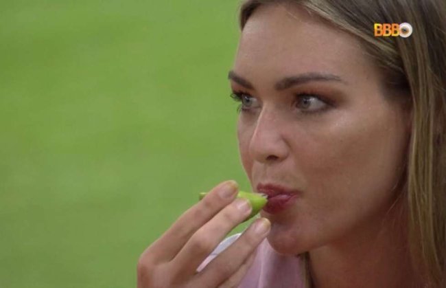 A participante do Big Brother Brasil, Bárbara Heck, está sentada no gramado, chupando a fruta limão. Ela é branca, dos olhos azuis e cabelos loiros.