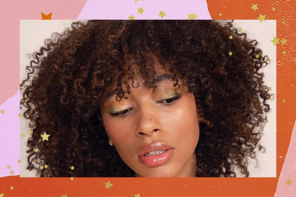 Montagem com o fundo colorido em tons de laranja e rosa com detalhe de estrelas douradas com a foto do close do rosto de uma mulher. Na foto ela está com o cabelo cacheado solto, maquiagem simples, olha para baixo e não sorri.