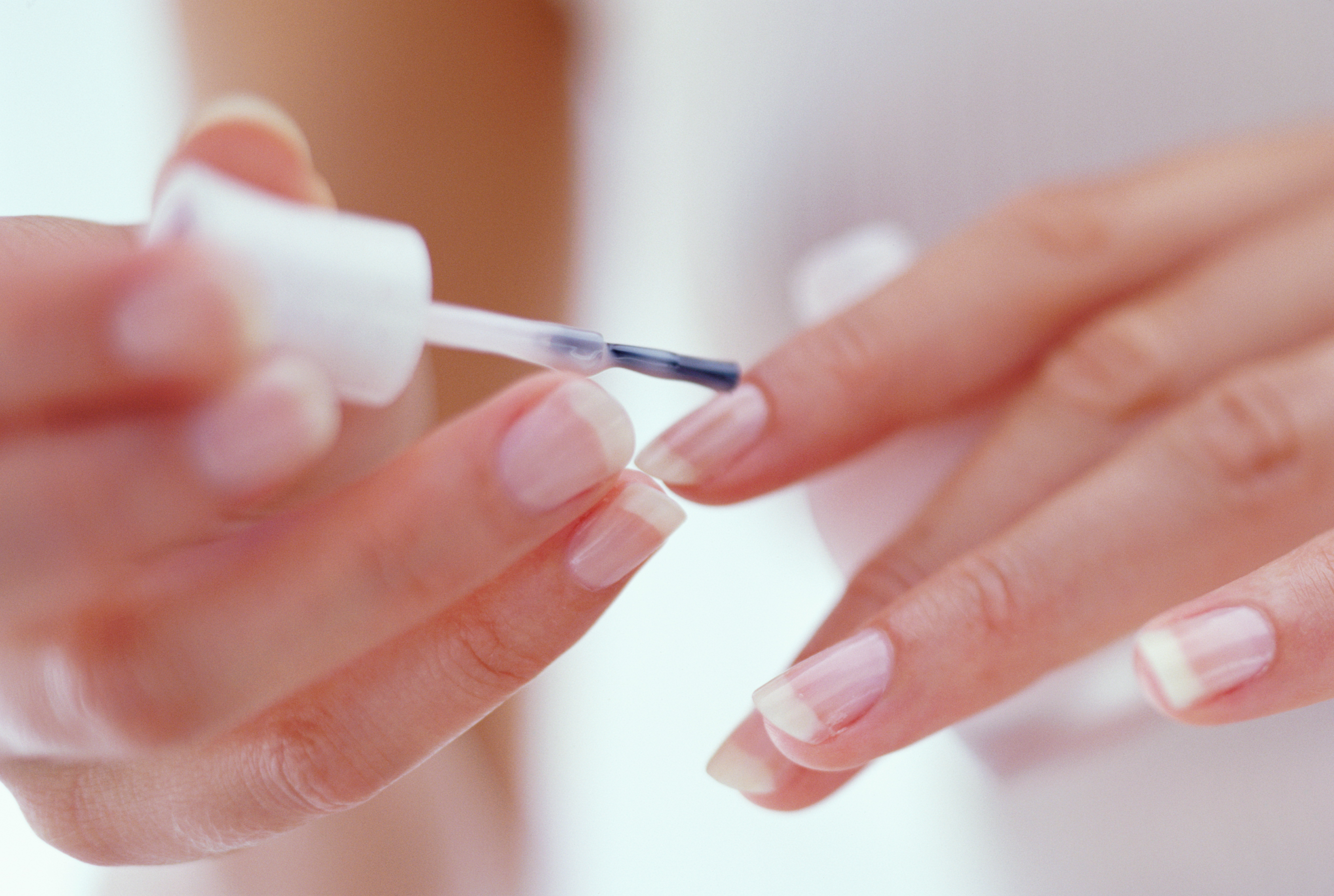 dicas-para-consertar-unha-em-casa Mulher jovem pintando as unhas com base transparente. A imagem destaca esse processo em close-up, plano da câmera que possibilita uma visão próxima e detalhada da fotográfica.