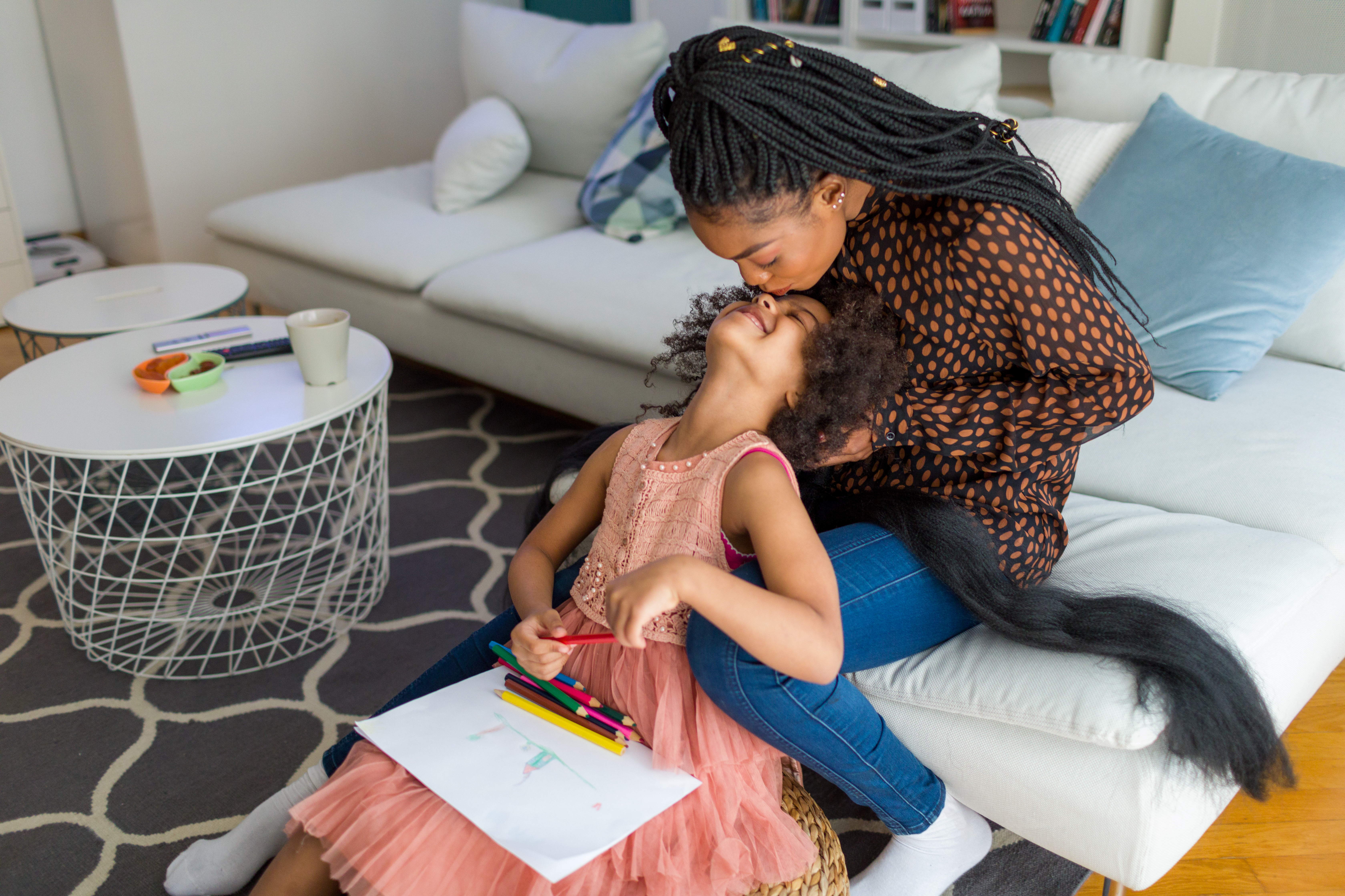 Imagem mostra mãe e filha sentadas no sofá e no chão, respectivamente. Ambas então em uma sala de estar enquanto a mãe trança o cabelo da filha e a mesma olha para cima e recebe um beijo na testa. O registro de trocas e afetos entre mãe e filha segue acompanhado de uma borda colorida e estrelinhas douradas por toda a imagem.