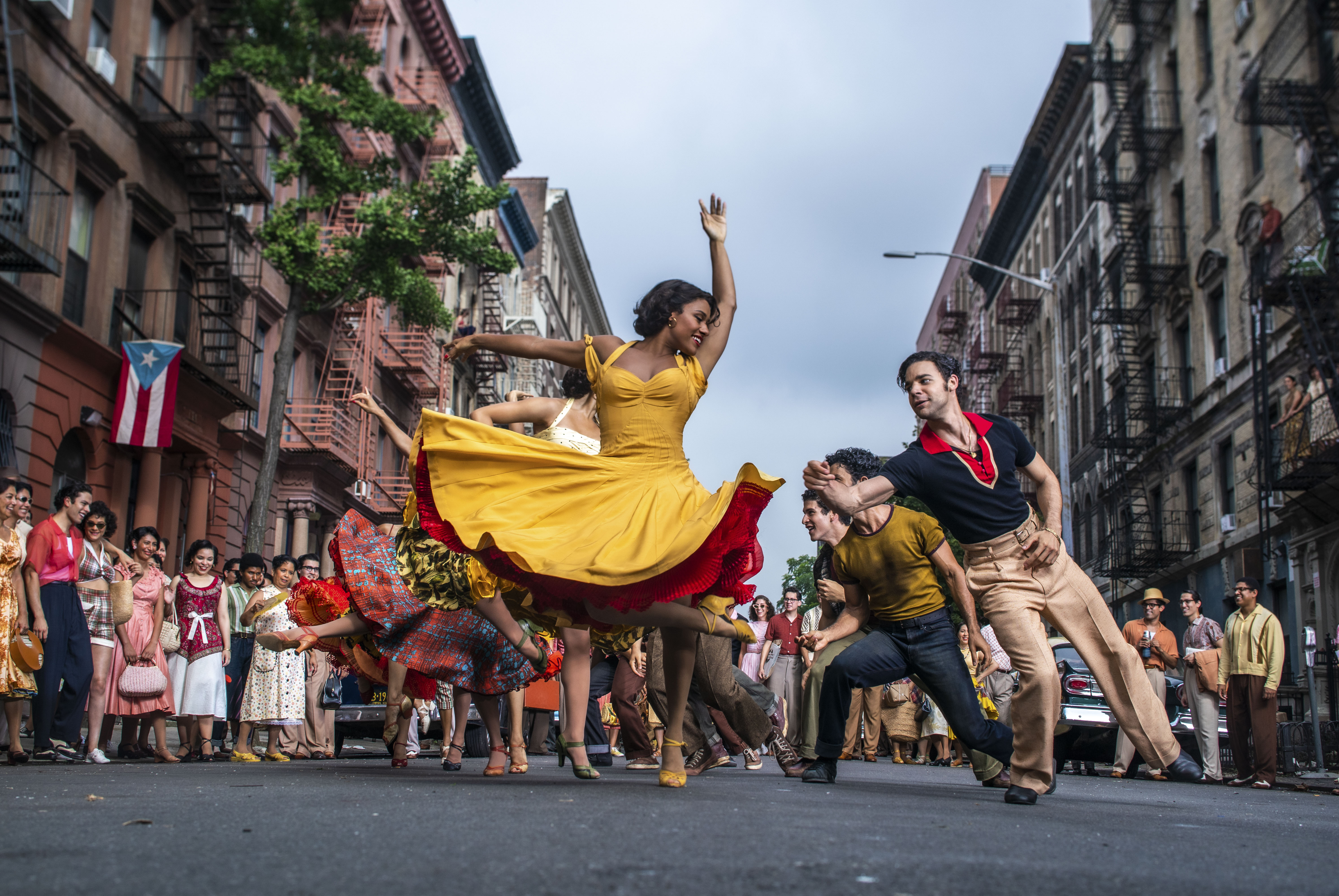 Ariana DeBose como Anita e David Alvarez como Bernardo em cena de Amor, Sublime Amor; eles estão dançando na rua com outras pessoas também dançando ao fundo; ela usa um vestido amarelo com babados vermelhos na saia e ele usa uma camisa azul com detalhes em vermelho