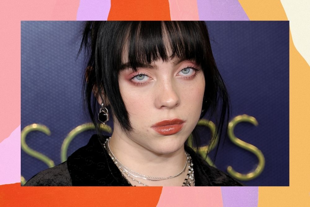 Billie Eilish posando para foto no tapete vermelho; a margem é uma textura nas cores rosa, lilás, laranja, roxo, amarelo e branco