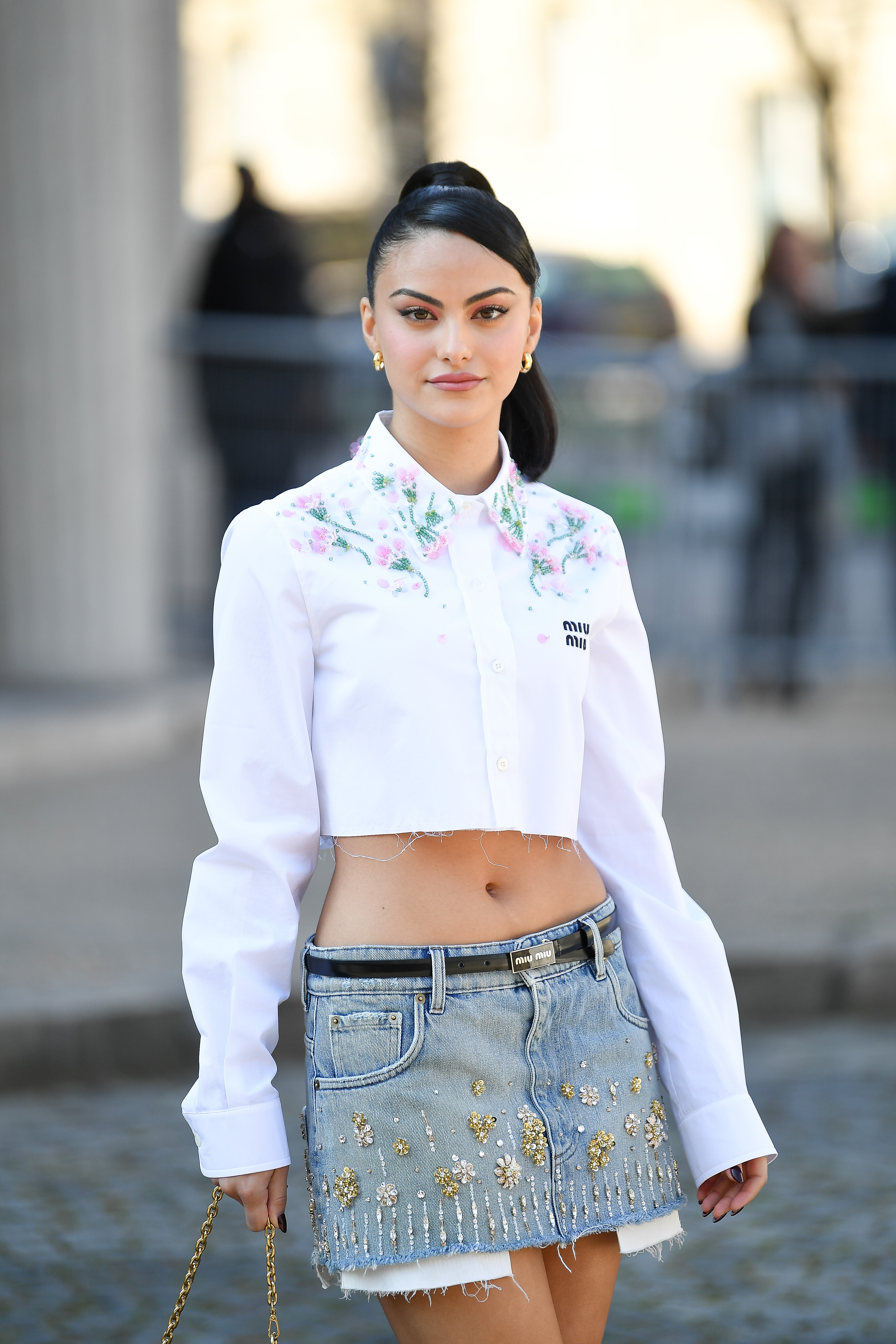 Camila Mendes usando camisa branca cropped, minissaia jeans e cabelo preso em rabo de cavalo alto