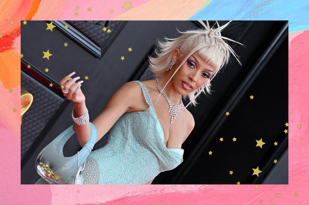 Montagem com o fundo colorido e detalhe de estrelas douradas com a foto da Doja Cat no Grammy 2022 no centro. Na foto ela usa um vestido azul, bolsa de vidro azul, cabelo loiro preso e maquiagem. Ela olha para o lado e não sorri para a foto.