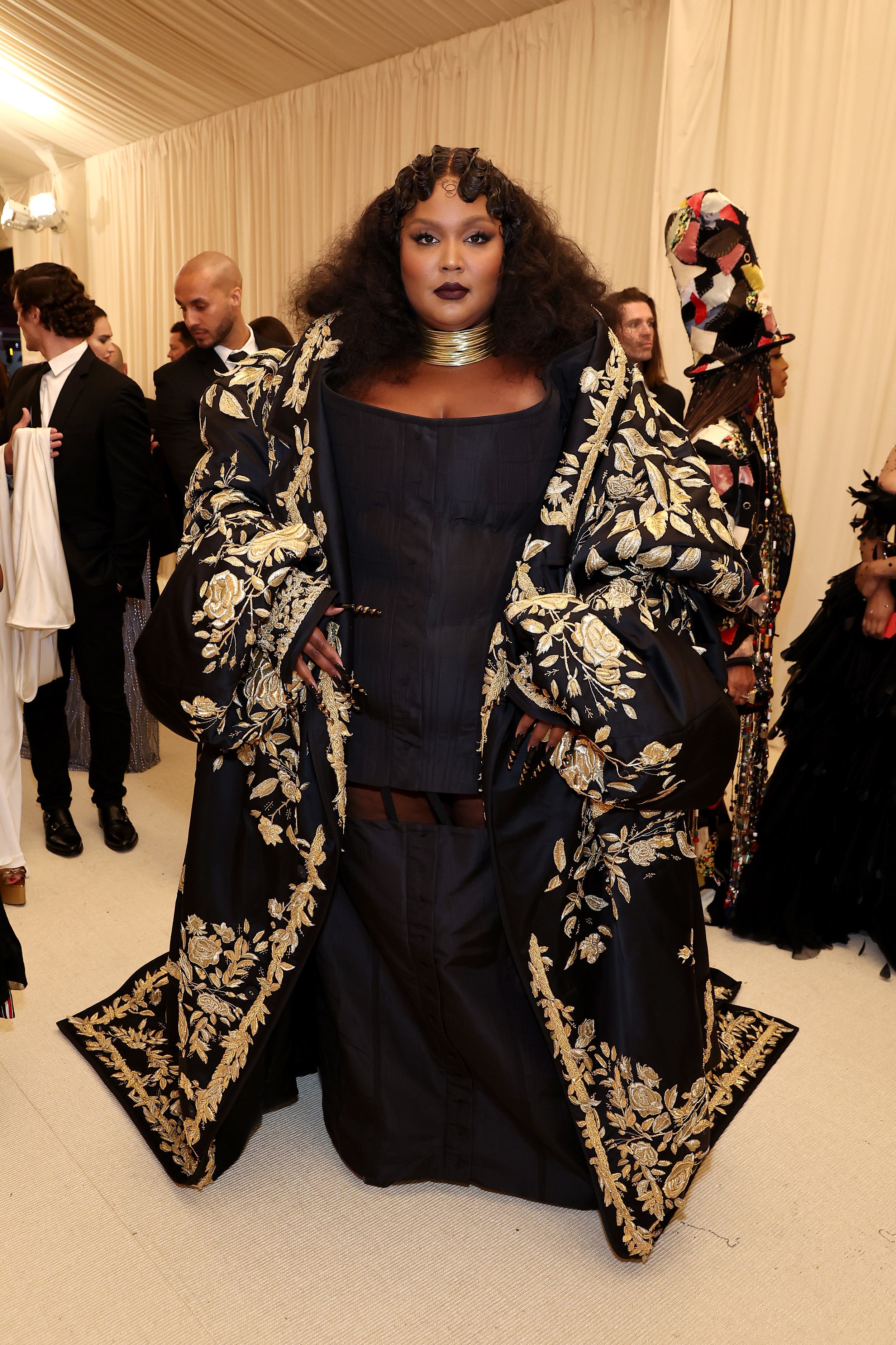 A imagem mostra Lizzo usando vestido preto e capa com bordado dourado no tapete vermelho do MET Gala 2022, no Metropolitan Museum of Art em 02 de maio de 2022 na cidade de Nova York.