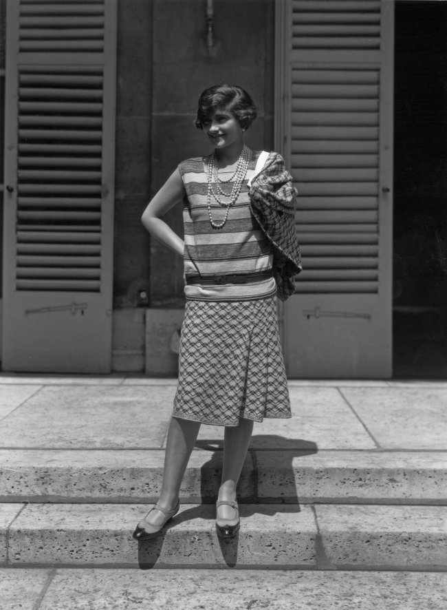 Foto em preto e branco da Coco Chanel de 1929. Ela usa uma camisa larguinha de listras, saia midi estampada e sapatos mary jane. Ela olha para o lado e sorri levemente.