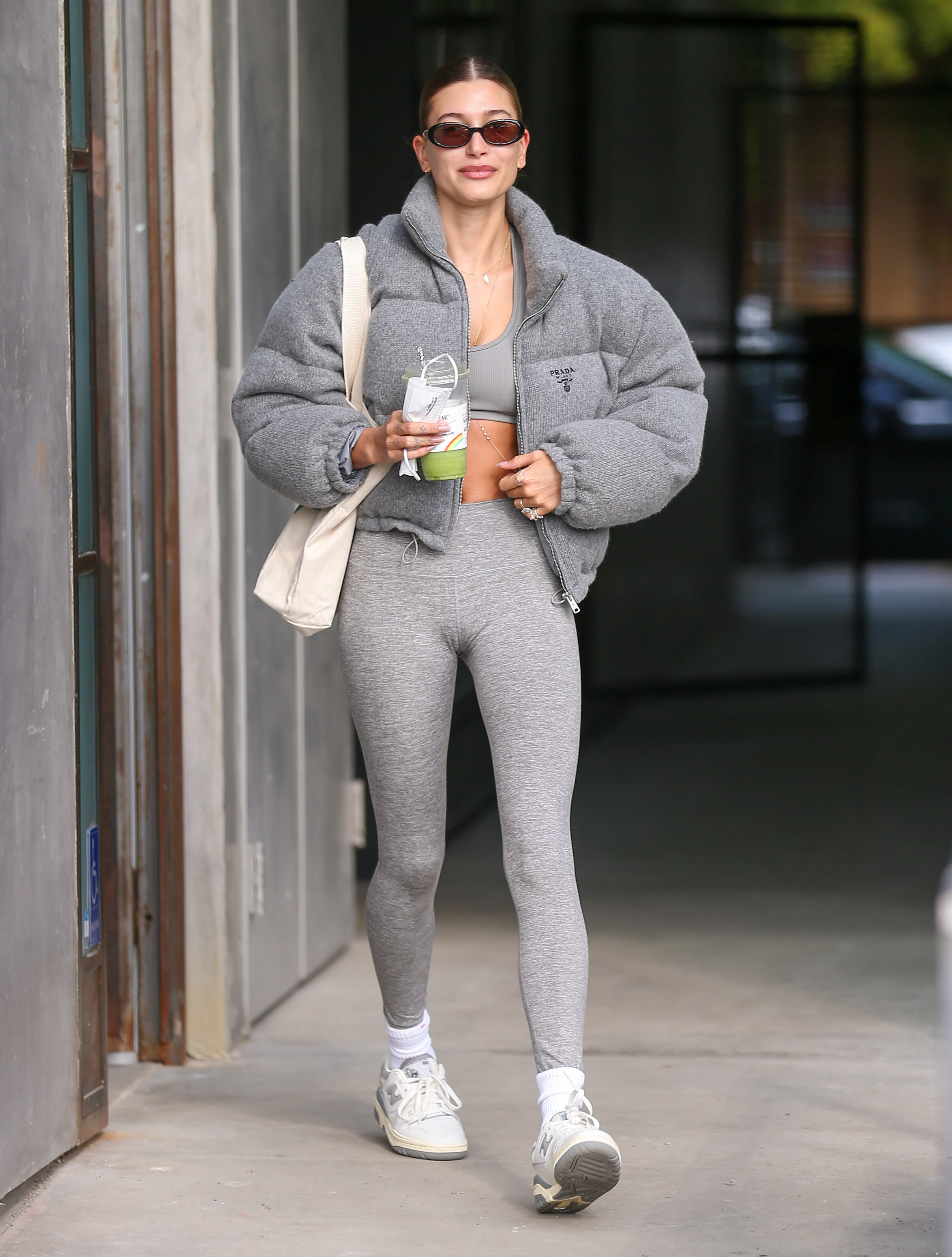 Hailey Bieber usando look todo cinza com jaqueta puffer, top, legging, tênis branco, com uma ecobag em um dos ombros e um óculos de sol