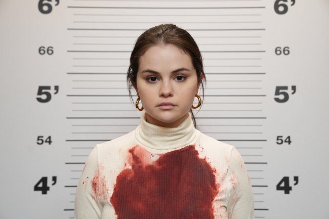 Only Murders In The Building Selena Gomez caracterizada como Mabel em Only Murders in The Building; ela está com expressão neutra posando para foto da polícia com medidores de altura ao fundo e sua blusa está manchada de sangue