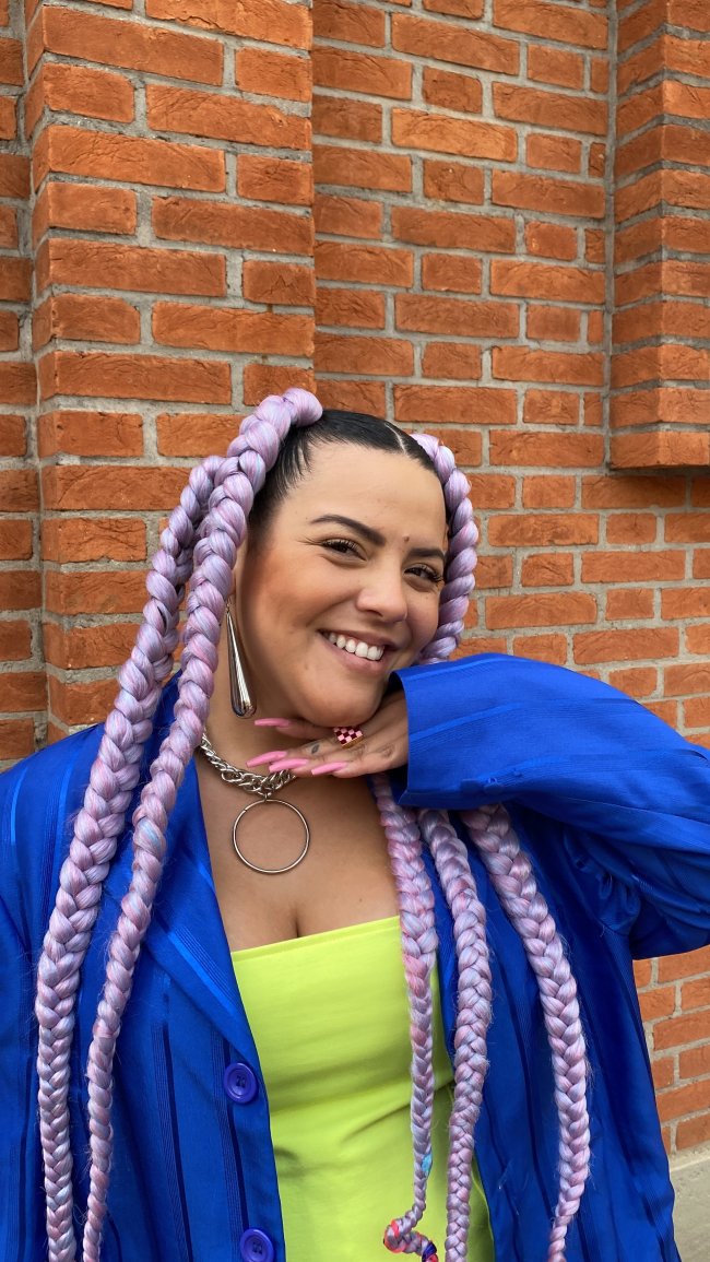 Imagem mostra Gab Alves sorrindo com a mão direita no queixo enquanto usa tranças e look colorido na spfw