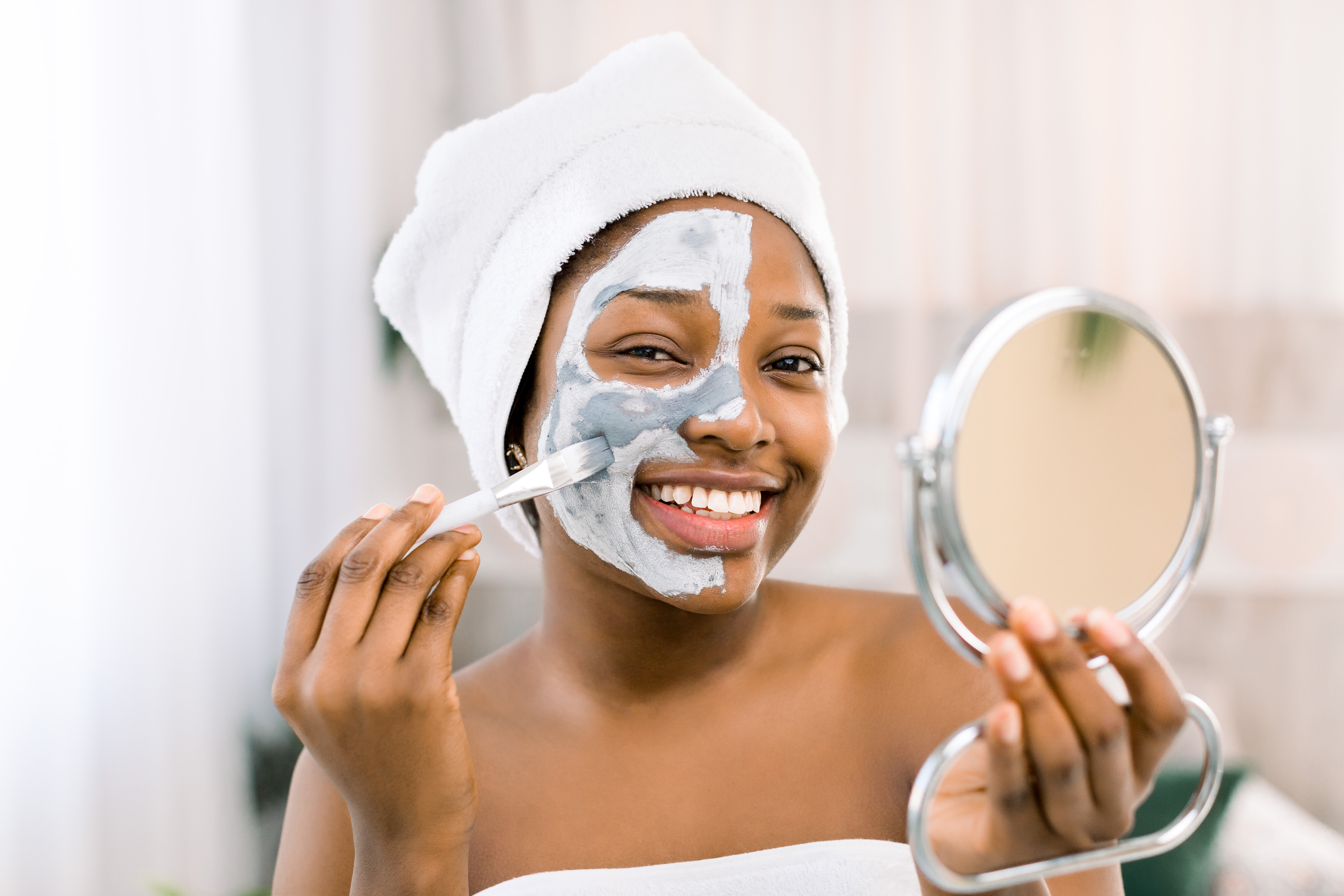 Garota sorrindo e fazendo skincare com máscara facial de argila com a ajuda de um pincel e um espelho