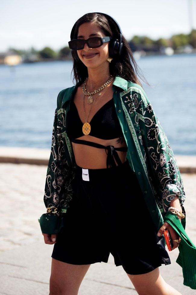 copenhagen-fashion-week-look-fone-de-ouvido Foto da Selma Kaci na Copenhagen Fashion Week. Na foto ela usa um top cropped preto, calça preto e casaco estampado. Ela está de cabelo solto, usa um fone de ouvido, olha e sorri para a foto.