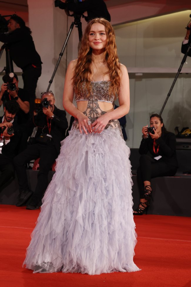 Foto da atriz Sadie Sink no tapete vermelho do Festival de Vezena. Ela usa um vestido com brilhos e recortes na parte superior e o restante do vestido com plumas. Ela está com o cabelo solto, maquiagem simples, olha para a câmera e sorri levemente.