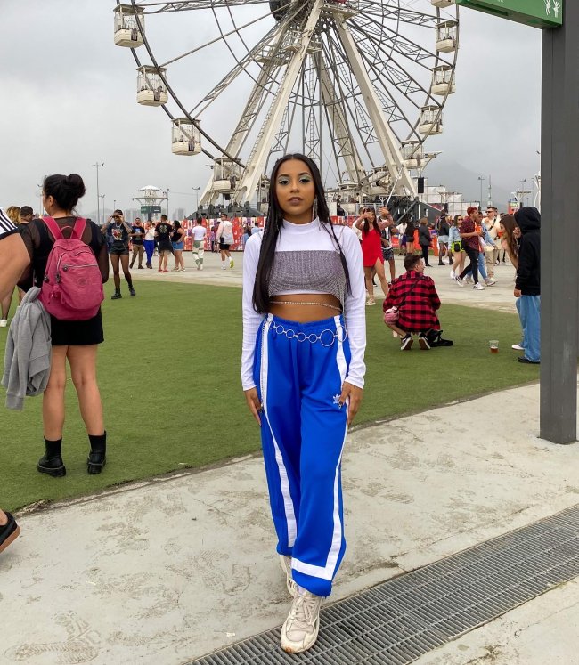 look-calca-azul-rock-in-rio-2022 Foto de uma menina no Rock in Rio 2022. Ela usa uma blusa de manga longa branca, top de brilhos como sobreposição e calça azul. Ela olha para a câmera e não sorri.