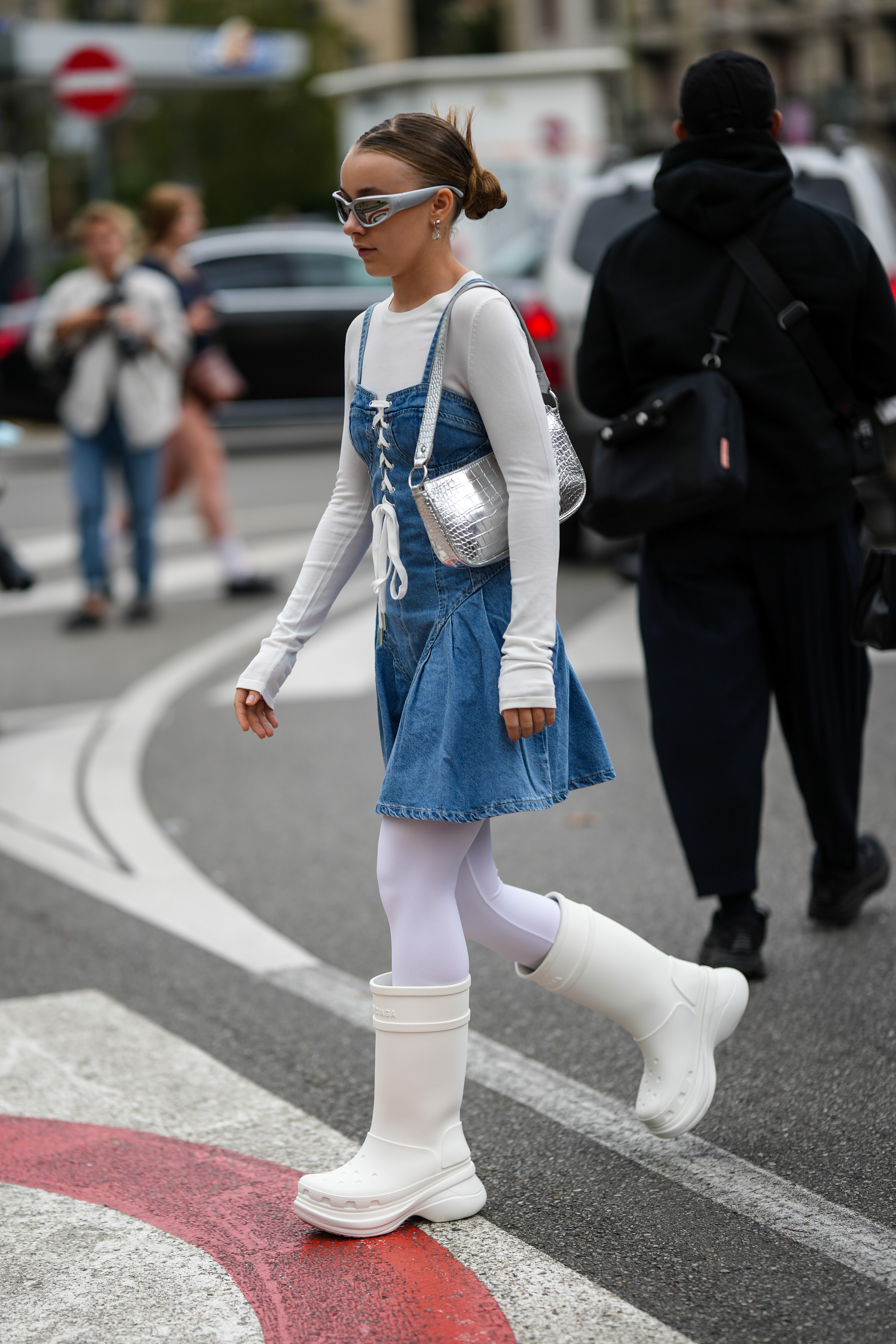 StreetStyle – Day 2 – Milan Fashion Week Womenswear Spring/Summer 2023 Garota usando vestido jeans por cima de blusa branca, meia-calça branca e galocha branca com óculos futurista e bolsa metalizada no street style da semana de moda de Milão