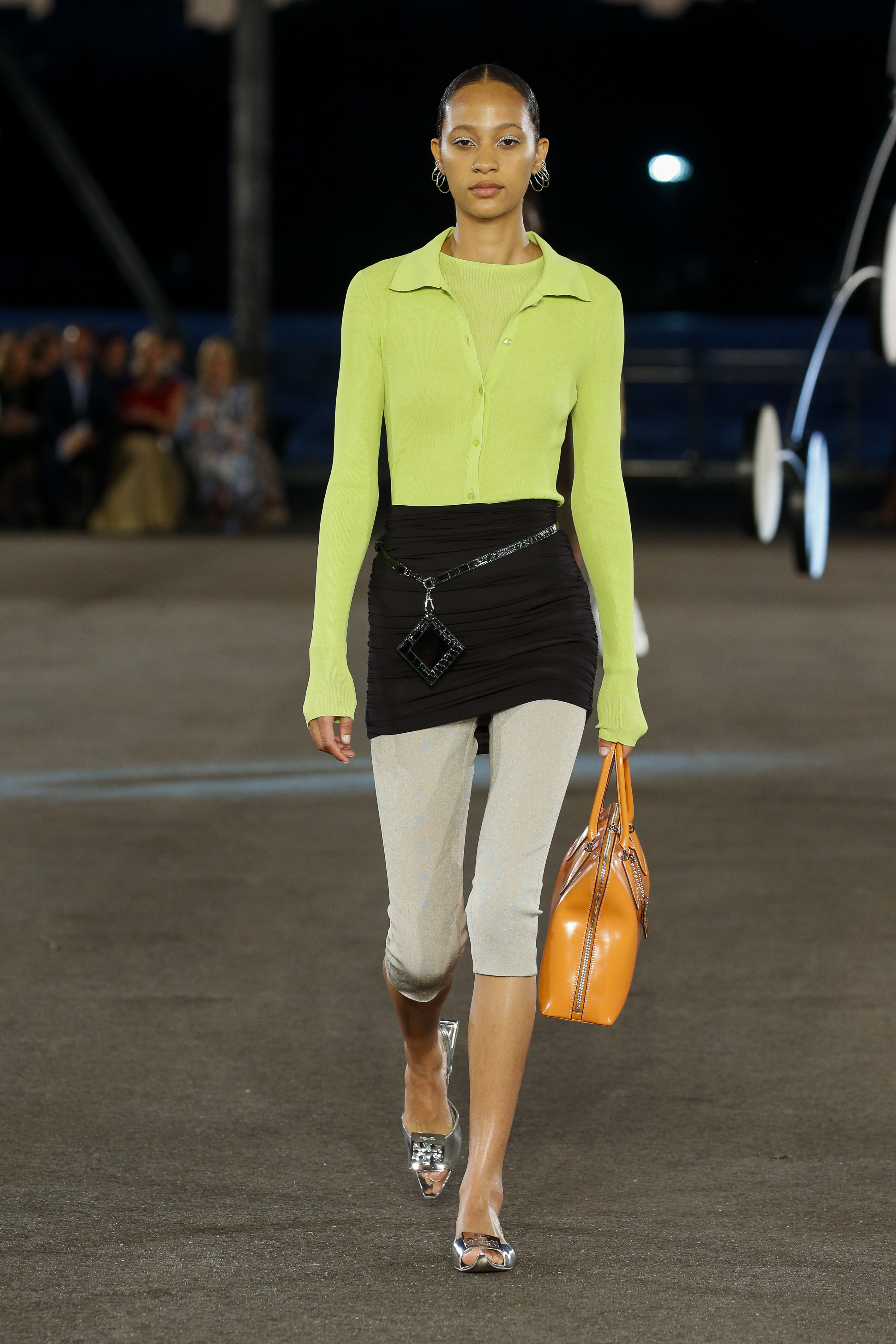 Tory Burch – Runway – Spring/Summer 2023 New York Fashion Week Desfile de primavera-verão 2023 da Tory Burch na semana de moda de Nova York. Modelo usando blusa de manga comprida com decote polo em verde neon, saia preta por cima de legging cropped off white e segurando bolsa laranja