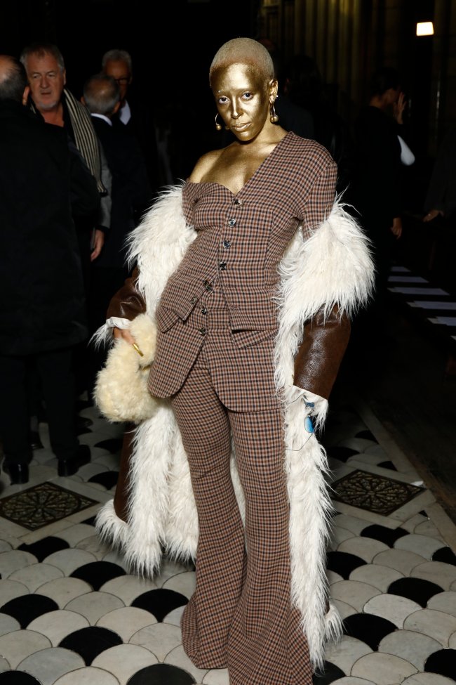 look-dourado-doja-cat-semana-de-moda-de-paris Foto da Doja Cat na Semana de Moda de Paris com um conjuntinho xadrez, casaco marrom de couro e rosto pintado de dourado