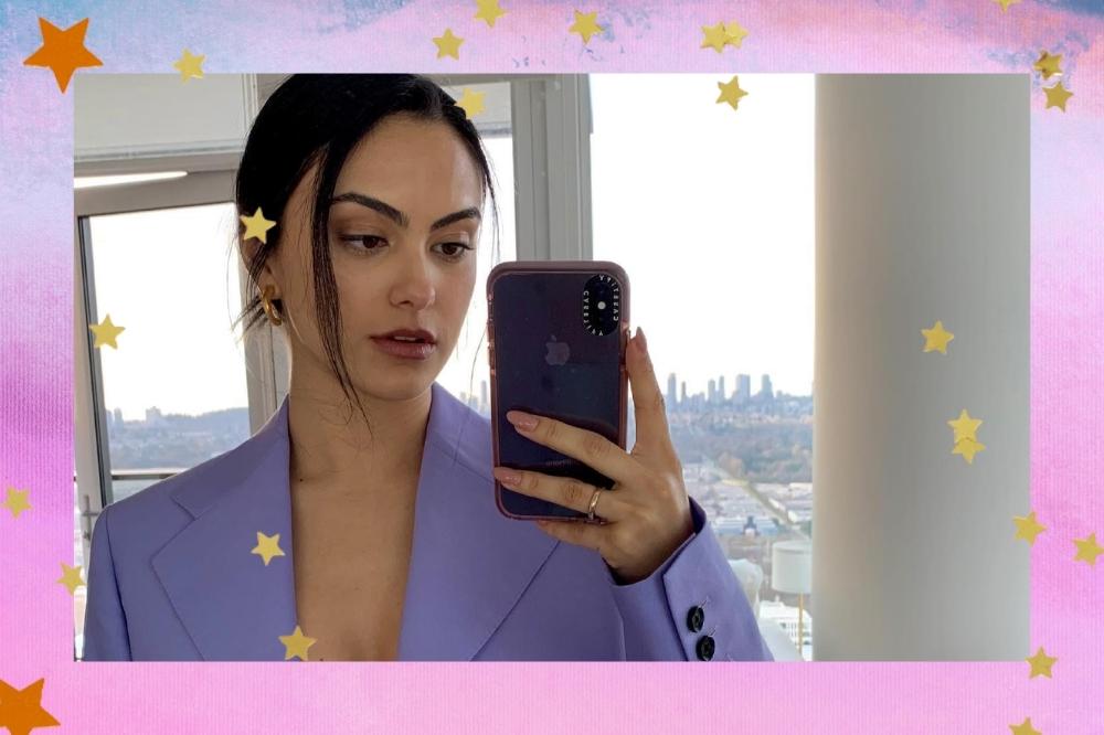 Montagem com o fundo rosa e detalhe de estrelas douradas nas bordas com uma selfie no espelho da atriz Camila Mendes no centro. Na foto ela usa um blazer lilás e cabelo preso em um coque baixo. Ela olha para o celular e não sorri.