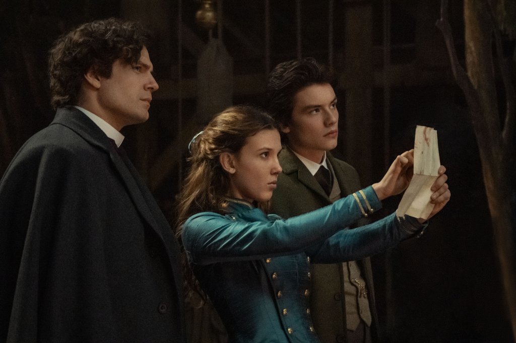 Henry Cavill como Sherlock Holmes, Millie Bobby Brown como Enola Holmes, Louis Partridge como Tewkesbury em imagem de Enola Holmes 2; eles estão olhando para um papel com atenção e expressões neutras e curiosas