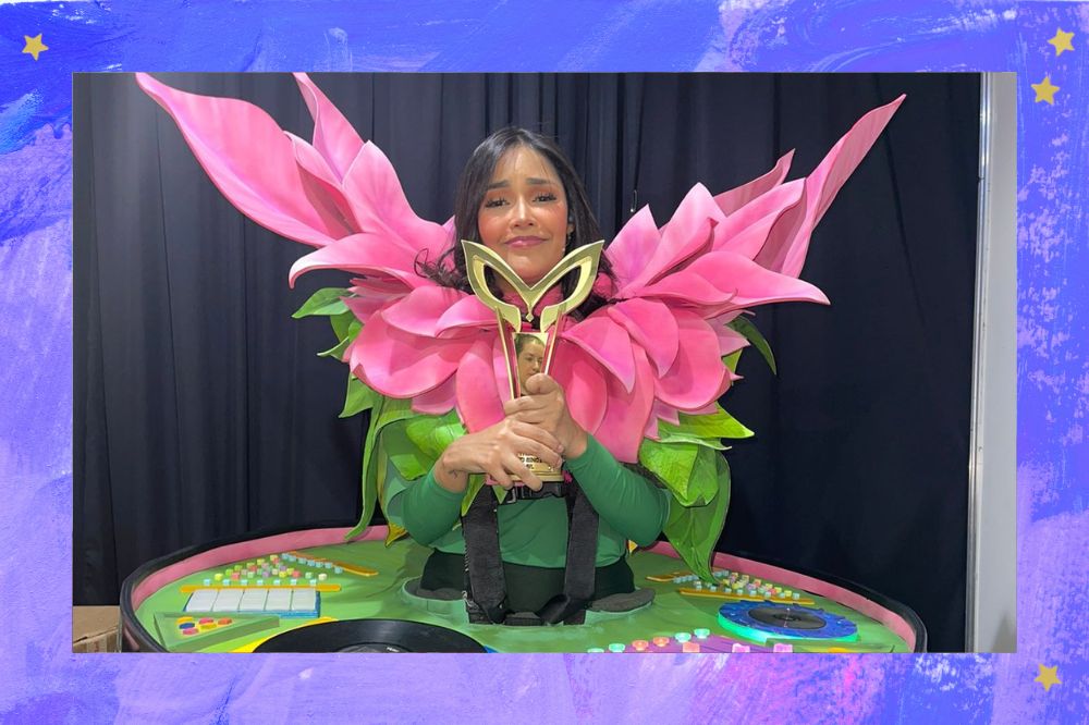 Flay caracterizada como personagem do segurando o troféu do The Masked Singer Brasil enquanto sorri levemente com expressão emocionada em foto; a margem é uma textura nas cores lilás, azul, roxo e branco; estrelas amarelas decoram a imagem