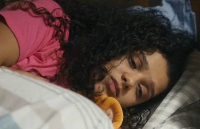 Karina, de Travessia, chorando na cama. Ela é uma menina negra de cabelos longos e cacheados