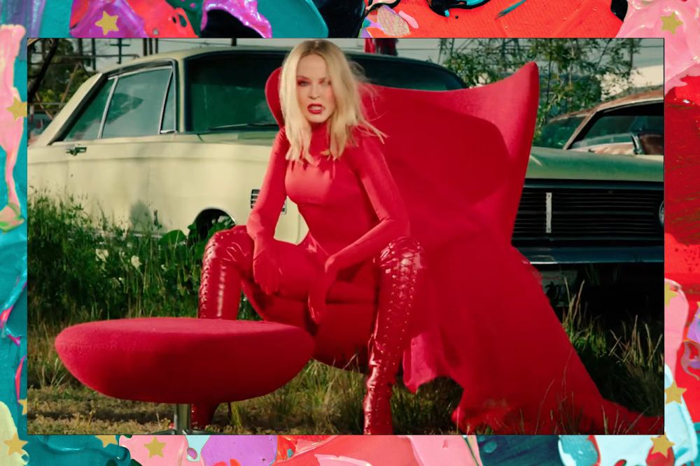 Kylie Minogue vestida de vermelho, sentada em uma poltrona vermelha do lado de fora de um trailer. Fundo colorido com tons de vermelho, rosa, lilás e azul.