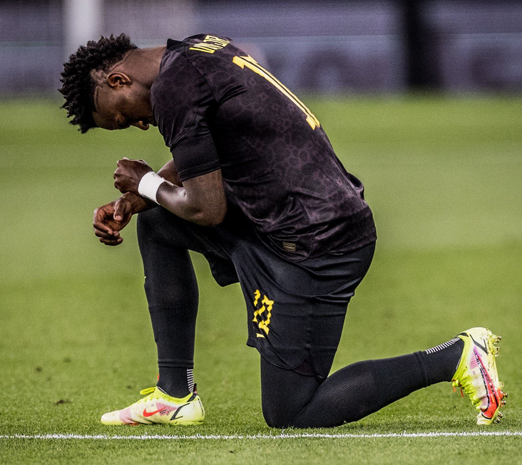 Vini Jr usa o uniforme preto da seleção brasileira. O jogador está ajoelhado no campo de futebol com uma das pernas a frente