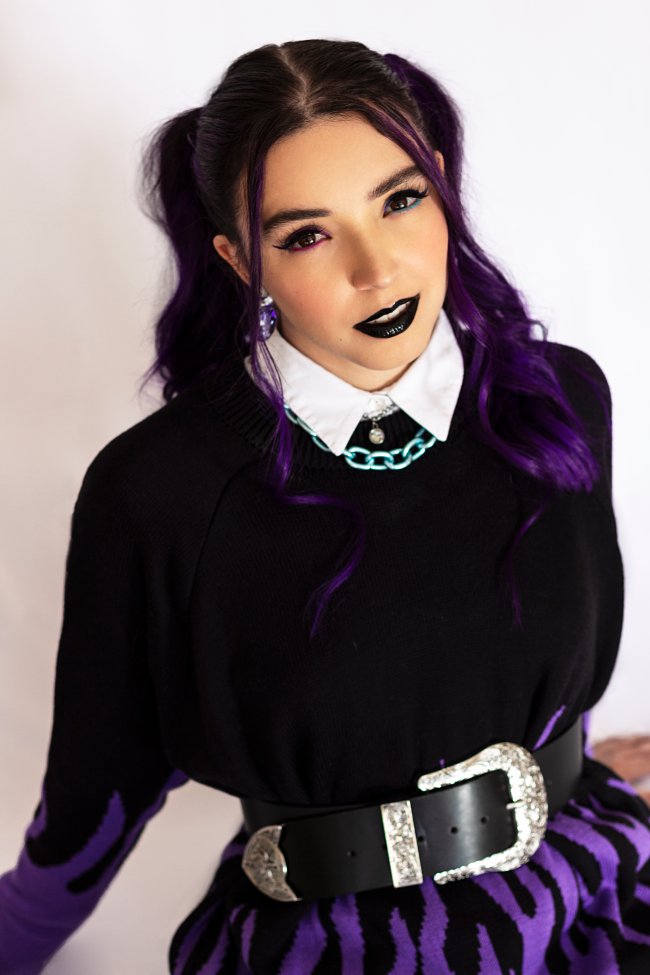 ana-xisde-maquiagem Ana Xisdê encara a cêmera enquanto usa batom preto e delineado. Seu cabelo roxo está preso em maria chiquinha e veste blusa de manga comprida preta com gola branca aparente, cinto grande com fvela prata e saia preta também.