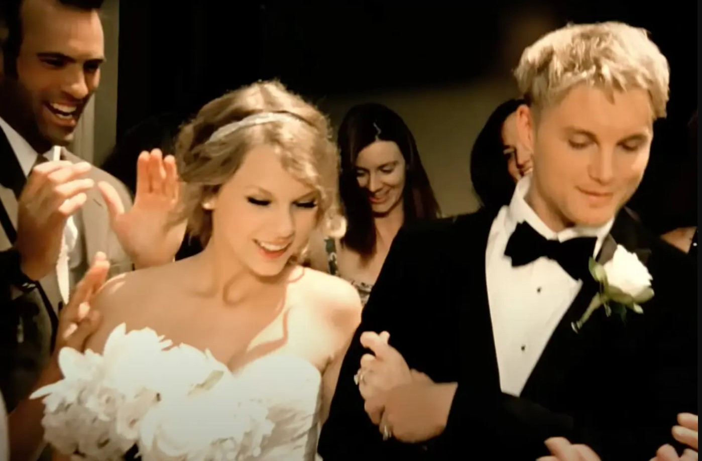 taylor-swift-mine Taylor Swift vestida de noiva com um buquê de flores brancas na mão e um vestido tomara que caia branco. Um homem louro - como um noivo - ao lado com um smooking preto. A cena é do clipe de Mine.