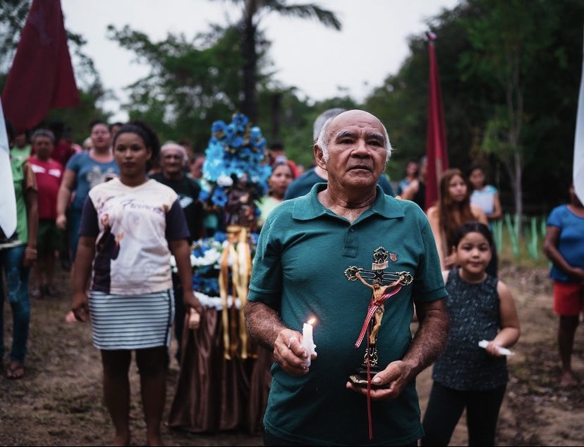 Um senhor de cabelos brancos, usando uma camisa verde, segura uma vela na mão esquerda e na outra, segura a imagem do santo