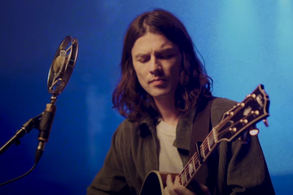 James Bay em pé tocando violão em um ambiente fechado com iluminação azul; ele está com expressão concentrada olhando para o instrumento