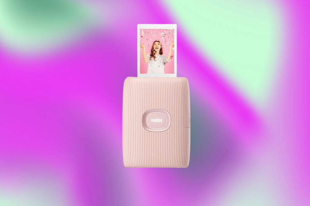 Impressora pequena na cor rosa claro da Instax. Dela está sendo revelada uma foto de uma mulher. O fundo da imagem é rosa, roxo e verde claro.