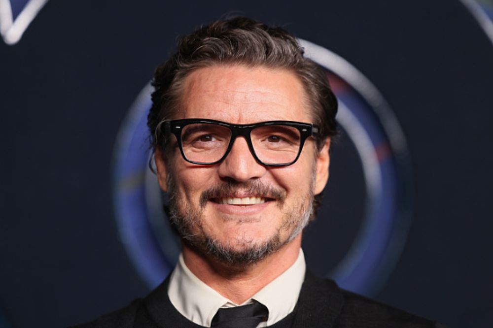 Foto do ator Pedro Pascal sorrindo para a câmera. Homem branco com cabelo penteado para trás, usando óculos azul, terno clássico.