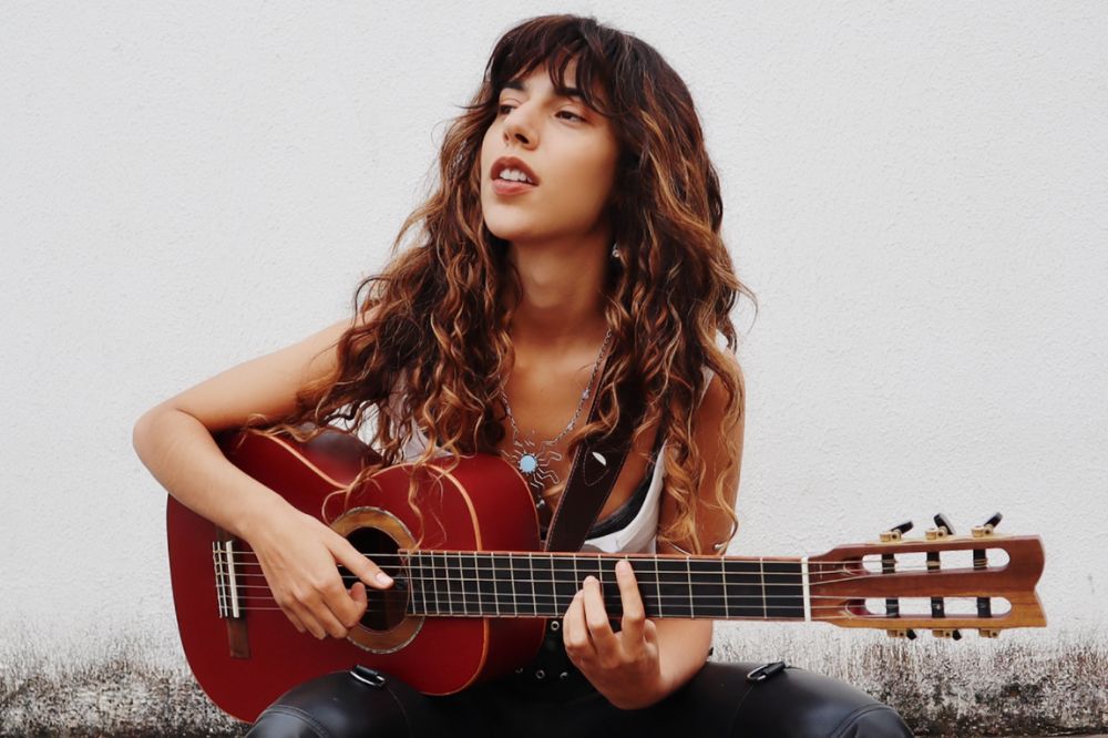 Mulher branca segurando violão vermelho. Cabelo ondulado castanho claro com franja.