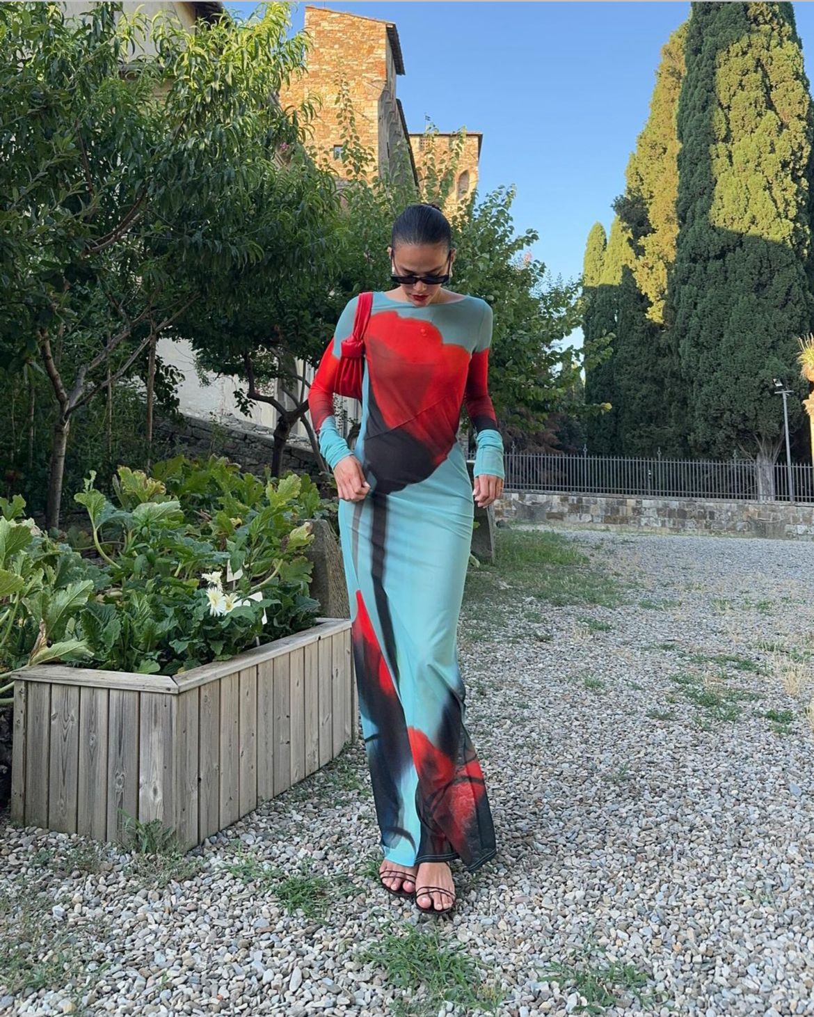 Imagem de corpo inteiro de Bruna Marquezine, mulher branca de cabelos pretos. Ela está olhando para baixo em um lugar com jardim, vestindo um vestido longo justo com transparência vermelho, preto e azul.