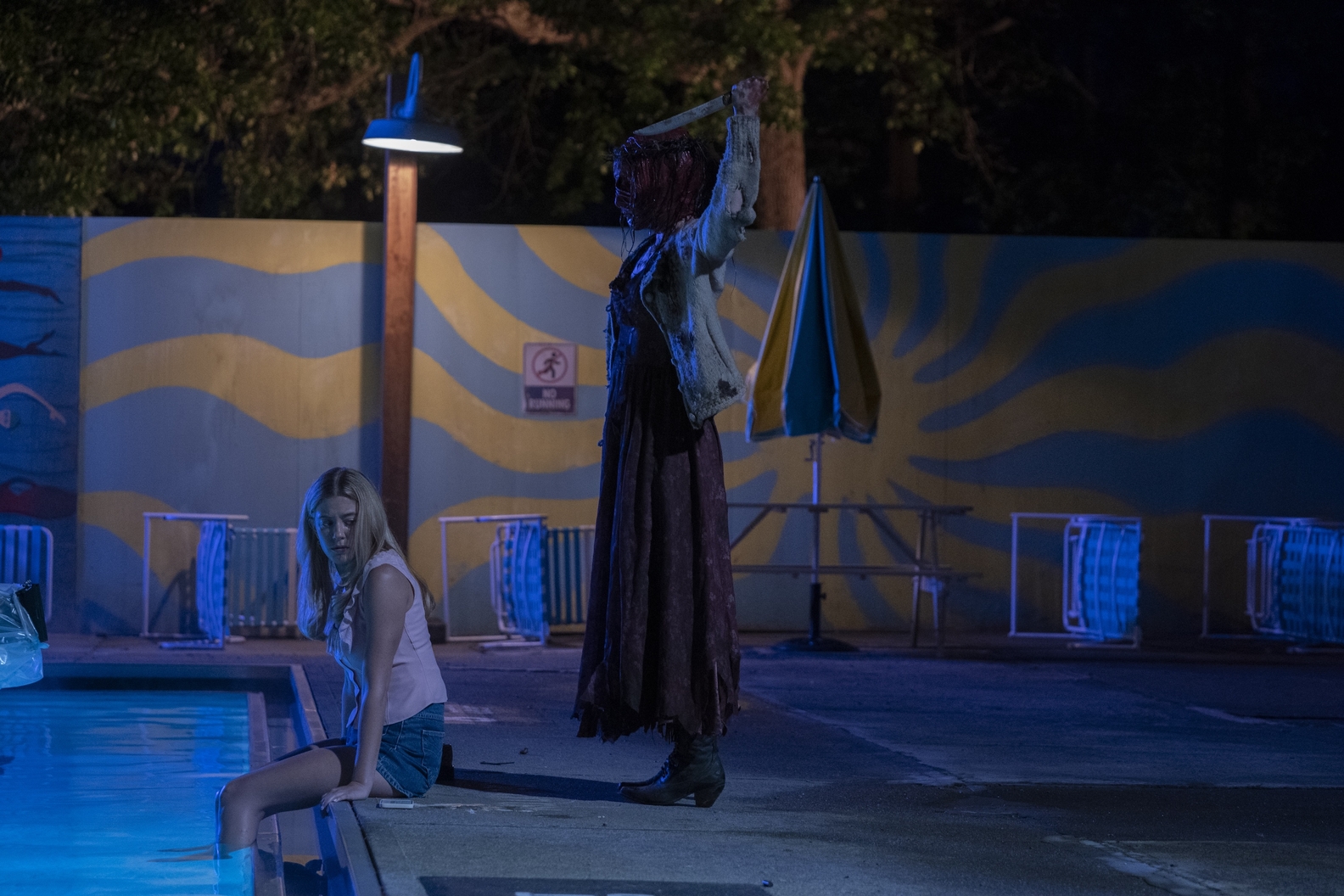 mallory-bechtel-bloody-rose_0 Bloody Rose, personagem de Pretty Little Liars: Summer School, com bandagens cobrindo o rosto e coberta de sangue enquanto levanta uma faca na direção de Karen, personagem que está sentada na beira da piscina de costas para vilã enquanto se vira